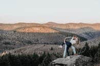 A couple embracing on a rocky cliff with a scenic mountain landscape in the background.