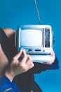 A woman holds a retro portable TV against a vibrant blue background, evoking nostalgia.