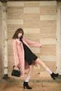 Fashion model in pink coat and black dress striking a dynamic pose indoors with stylish black heels.