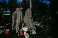 Unrecognizable children standing on dark street with buckets for trick or treat covered with white blankets painted like ghosts for Halloween celebration