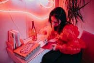 A woman writes a holiday card in a vibrant, neon-lit room with a festive atmosphere.