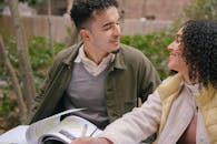 Smiling young Hispanic couple in warm clothes sitting at table while preparing for exams with textbook near green shrubs and trees in daylight in park while looking at each other
