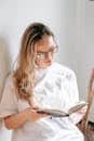 A young woman wearing eyeglasses attentively reading a book indoors in natural light.
