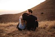 A couple sits closely together on a hill, sharing a warm embrace during a beautiful sunset in California.