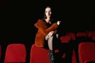 A woman in a fur jacket sits in a dimly lit movie theater with red seats.