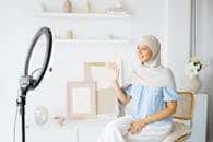 Smiling woman in hijab with ring light waving in a modern indoor setting.
