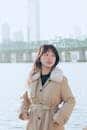 A stylish woman in a beige trench coat poses by the Seoul waterfront, showcasing urban fashion.