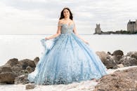 A woman in a blue gown poses by the sea with a castle in the background. Captures elegance and beauty by the shore.