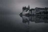 A somber castle reflects on a calm lake, shrouded in mist, offering a moody and serene atmosphere.