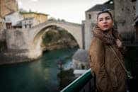 Fashionable woman in fur coat posing near a historic stone bridge during the day.