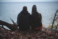 A couple sits on a cliff's edge overlooking the tranquil ocean, surrounded by fallen leaves.