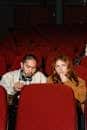 A couple seated in red cinema chairs enjoys a movie, sharing fun snacks.