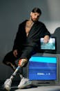 Man in a bathrobe with prosthetic legs sitting on CRT TVs in a conceptual studio setup.