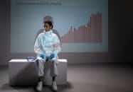 A healthcare worker in PPE sits by COVID-19 data chart. Illustrates pandemic impact.