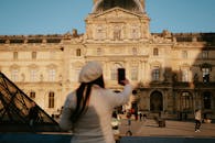 A woman with a smartphone takes a picture of the iconic Louvre Museum in Paris, France.