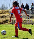 A girl in a red jersey playing soccer outdoors on grass with spectators in background.