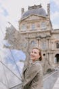 A cheerful woman poses in front of the iconic Louvre Museum in Paris, showcasing its glass pyramid.