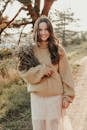 Smiling woman in cozy sweater holding a bouquet of dry flowers on a sunny outdoor path.