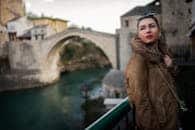 Fashionable woman in fur coat posing near a historic stone bridge during the day.