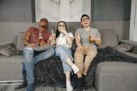 Three friends relaxing on a sofa with drinks, watching a movie and laughing indoors.
