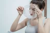 Woman in a tank top applying a moisturizing sheet mask for skincare treatment.