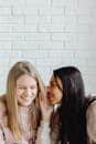 Two teenage girls whispering and smiling against a white brick wall, showcasing friendship and youthful joy.