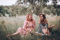 Two women enjoy a summer picnic in a scenic grass field, dressed in floral dresses, embodying friendship and style.